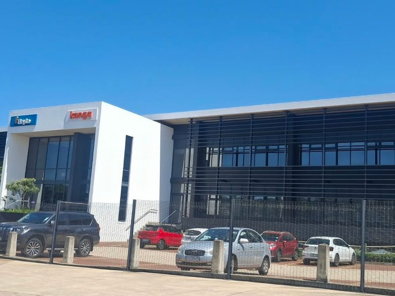 Modern commercial office building featuring a glass facade with metal louvers, secure perimeter fencing, and an adjacent paved parking area.