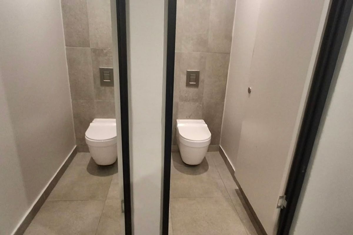 A modern commercial bathroom featuring two private stalls with wall-mounted toilets, concealed cisterns, and neutral tiled finishes.