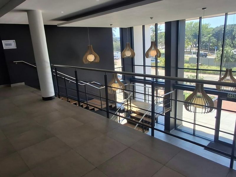 Modern commercial hallway featuring floor-to-ceiling glass windows, tiled flooring, and a sleek stainless steel balustrade overlooking the entrance area.