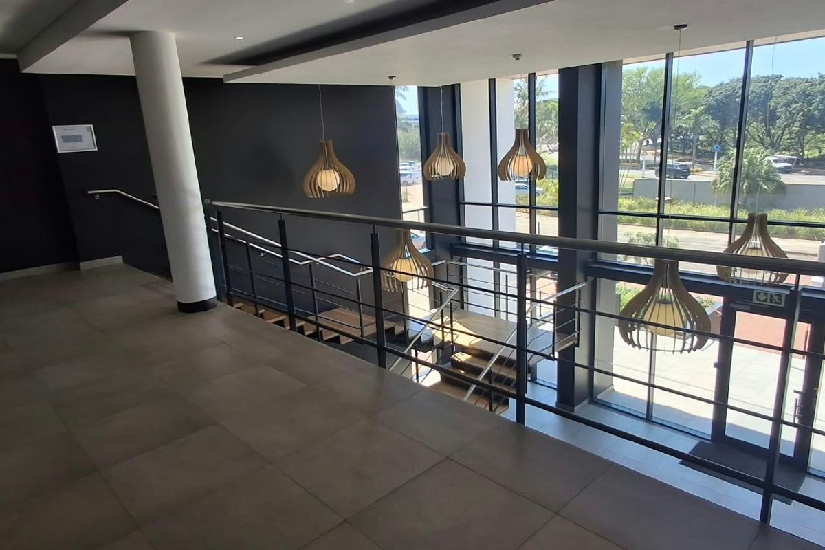 Modern commercial hallway featuring floor-to-ceiling glass windows, tiled flooring, and a sleek stainless steel balustrade overlooking the entrance area.