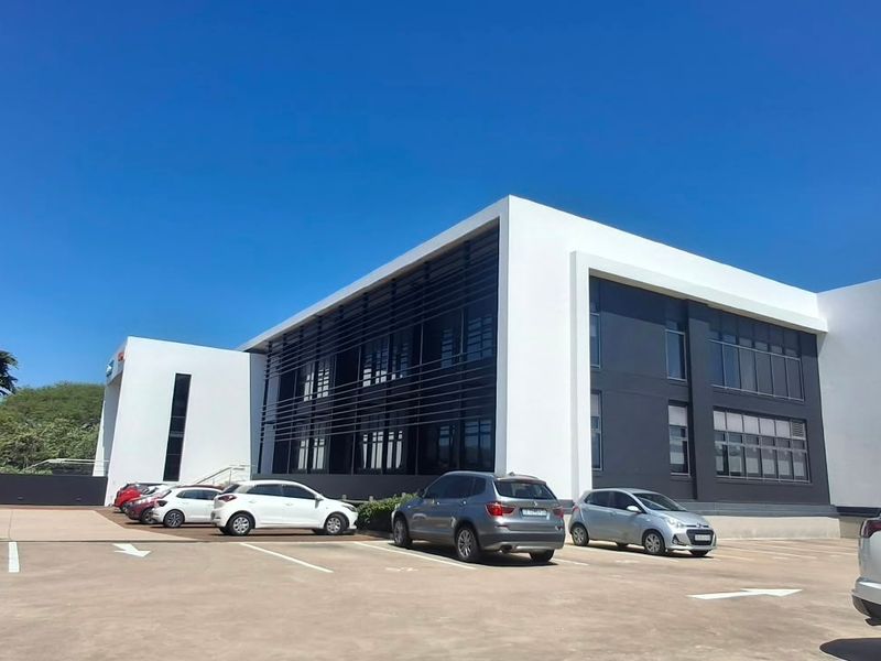 A modern commercial office building featuring a clean white and dark grey facade with extensive glass windows and an adjacent paved parking area.