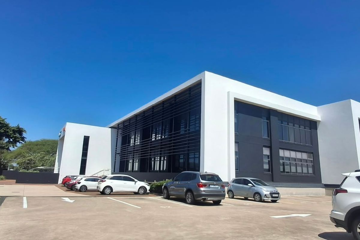 A modern commercial office building featuring a clean white and dark grey facade with extensive glass windows and an adjacent paved parking area.