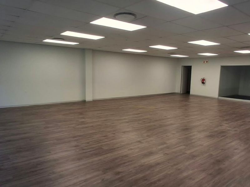 A spacious, open-plan office area featuring modern laminate flooring, a suspended ceiling with recessed lighting, and a glass-partitioned section.