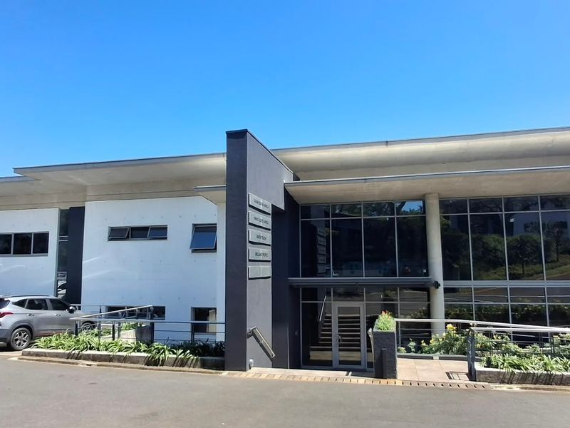 Modern office building exterior featuring a glass facade, concrete roof structure, and paved parking area with integrated landscaping.