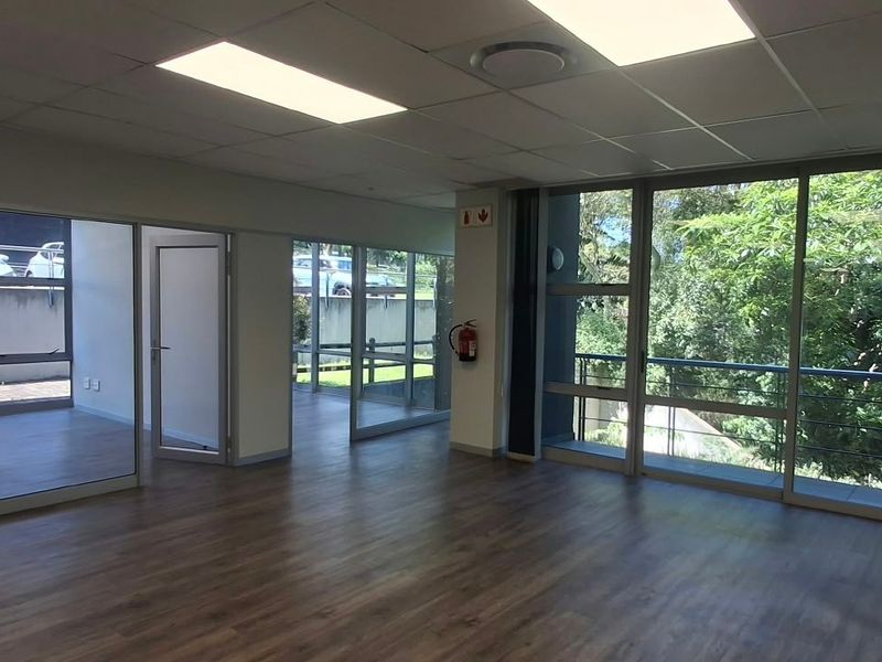 A bright, open-plan office space featuring modern laminate flooring, suspended ceilings with recessed lighting, and extensive glass partitioning providing natural light and balcony access.