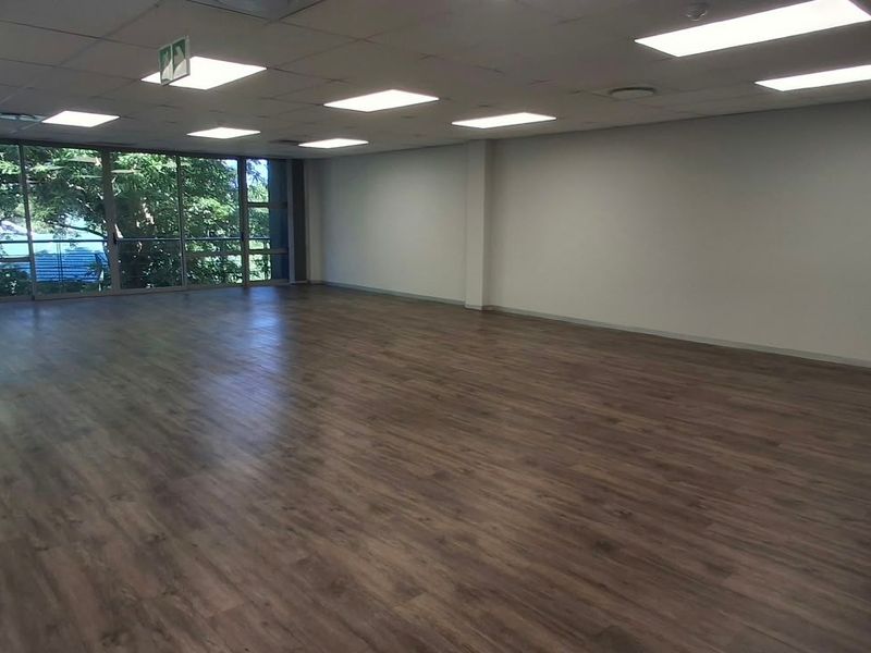 A spacious, open-plan office area featuring modern laminate flooring, a suspended ceiling with recessed lighting, and large windows providing natural light.
