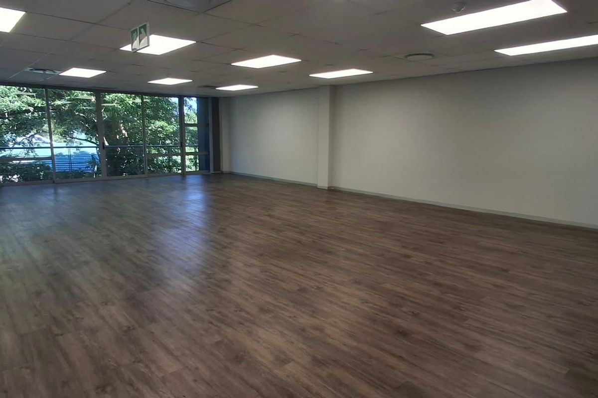 A spacious, open-plan office area featuring modern laminate flooring, a suspended ceiling with recessed lighting, and large windows providing natural light.