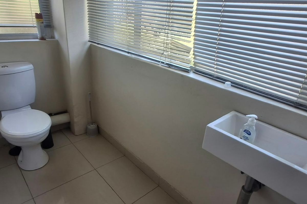 A functional bathroom featuring a white toilet, wall-mounted basin with a modern mixer tap, tiled flooring, and venetian blinds for privacy.