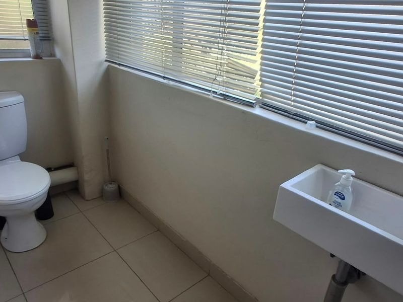 A functional bathroom featuring a white toilet, wall-mounted basin with a modern mixer tap, tiled flooring, and venetian blinds for privacy.