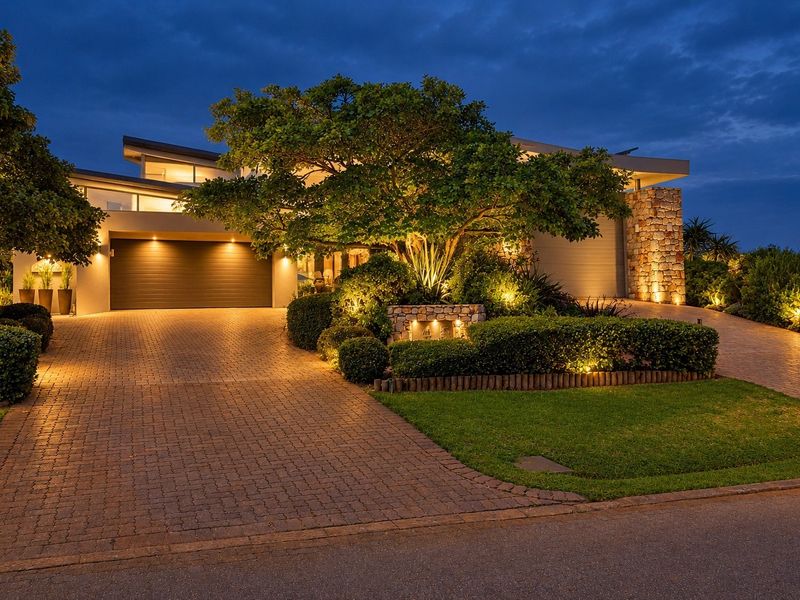 A beautifully illuminated modern residence featuring a paved driveway, double garage, and elegant stone-clad architectural details set within a manicured garden.