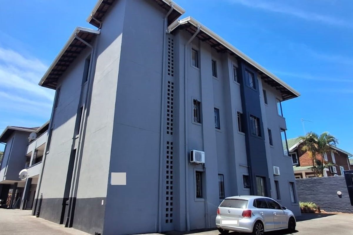 A multi-story residential building exterior featuring a modern grey facade, tiled roof, and paved driveway access.