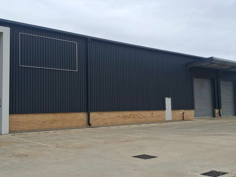 Industrial warehouse exterior featuring corrugated metal cladding, brick base, multiple roller shutter doors, and a paved concrete apron with marked parking bays.