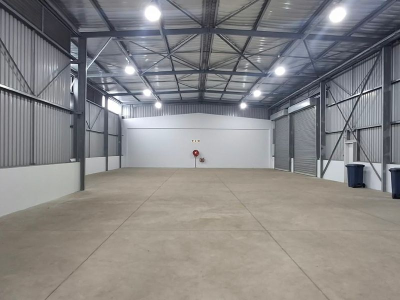 Spacious industrial warehouse unit featuring high ceilings with exposed steel trusses, concrete flooring, and large roller shutter doors for easy access.