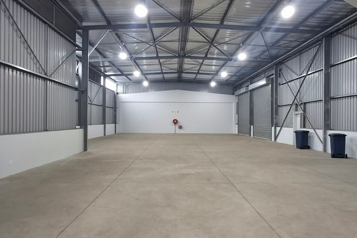 Spacious industrial warehouse unit featuring high ceilings with exposed steel trusses, concrete flooring, and large roller shutter doors for easy access.