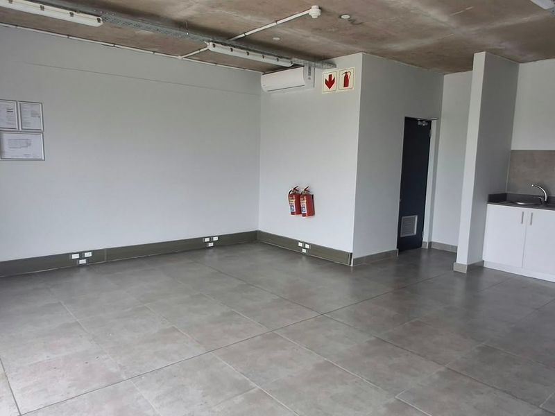 Open-plan industrial space featuring exposed concrete ceilings, tiled flooring, a compact kitchenette with built-in cabinetry, and essential fire safety infrastructure.