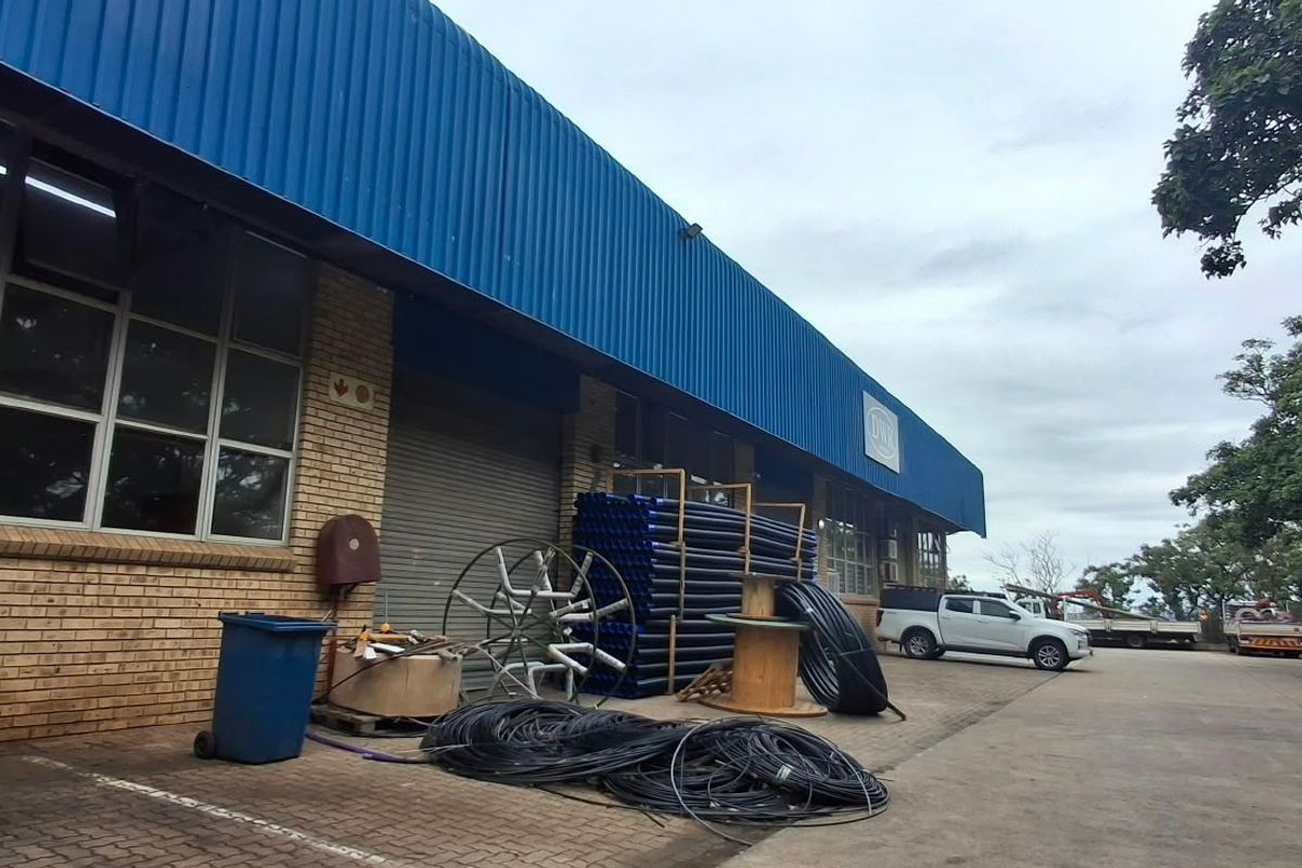 Industrial warehouse exterior featuring a blue corrugated metal roof, brick facade, and a roller shutter door with adjacent paved yard space.