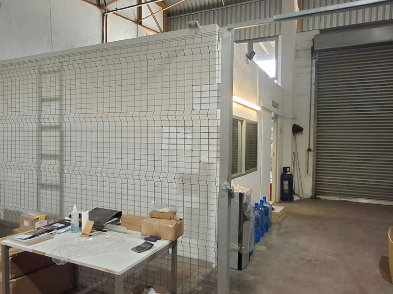 Industrial warehouse space featuring high ceilings with exposed steel trusses, a large roller shutter door, and secure wire mesh partitioning for storage or office areas.