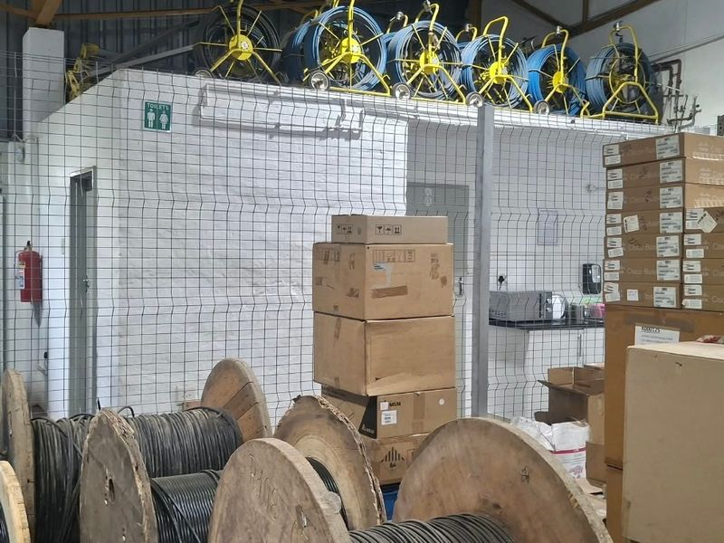 Industrial utility space featuring wire mesh partitioning, exposed steel roof trusses, and painted brick walls, currently utilized for storage and equipment housing.