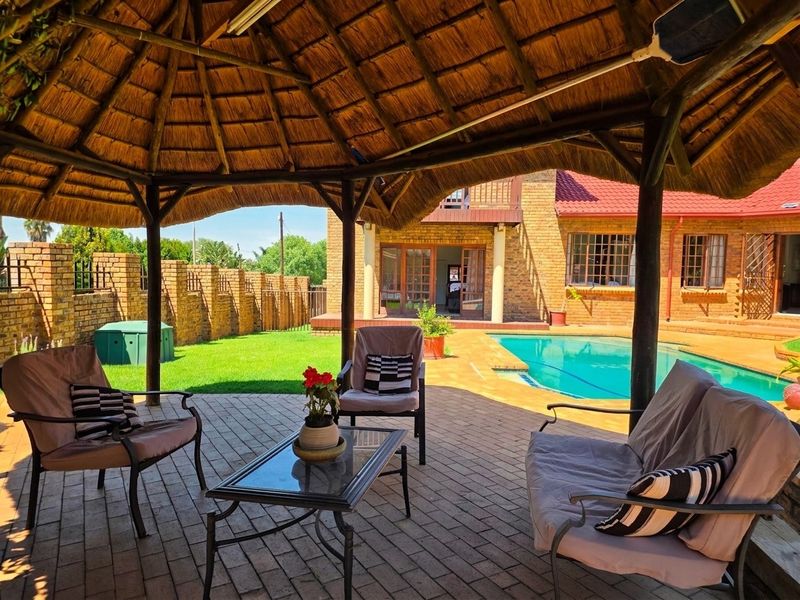 Covered outdoor patio area with a traditional thatch roof, overlooking a swimming pool and landscaped garden with brick boundary walls.