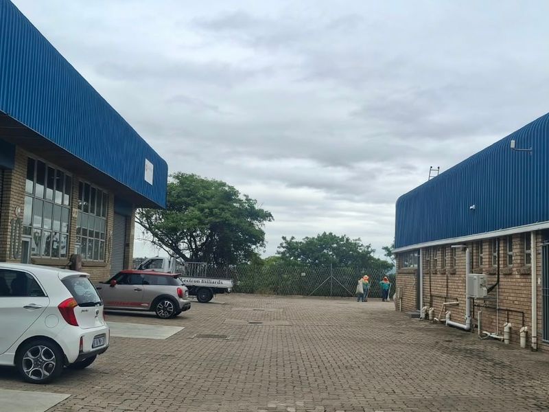 Exterior view of an industrial property featuring brick facades, blue corrugated metal roofing, roller shutter doors, and a paved yard area.