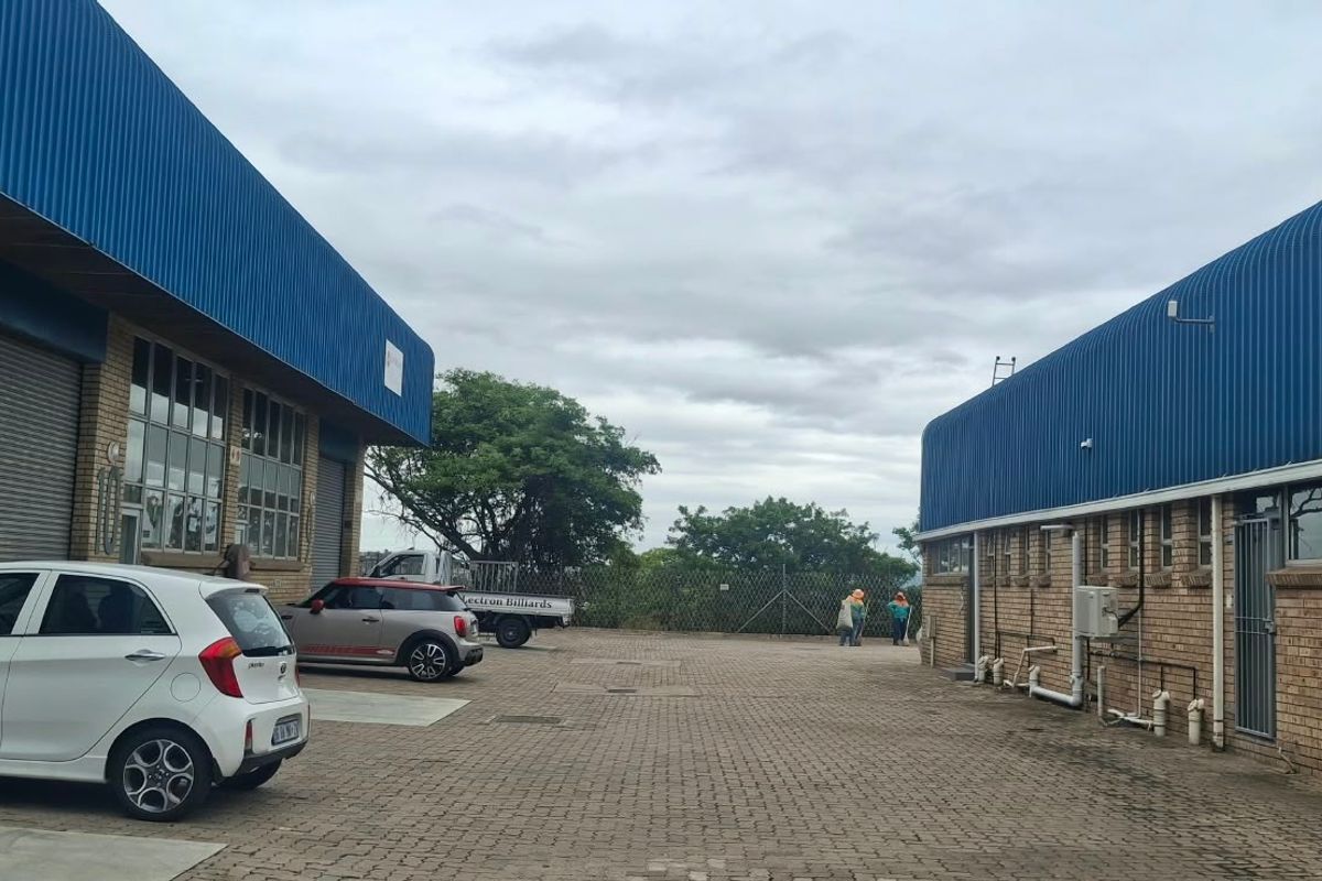 Exterior view of an industrial property featuring brick facades, blue corrugated metal roofing, roller shutter doors, and a paved yard area.