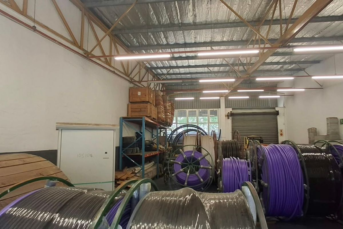 Industrial warehouse space featuring high ceilings with exposed steel trusses, roller shutter door access, and functional fluorescent lighting.