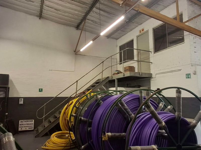 Industrial warehouse space featuring a mezzanine level with metal staircase access, exposed steel roof trusses, and functional overhead lighting.
