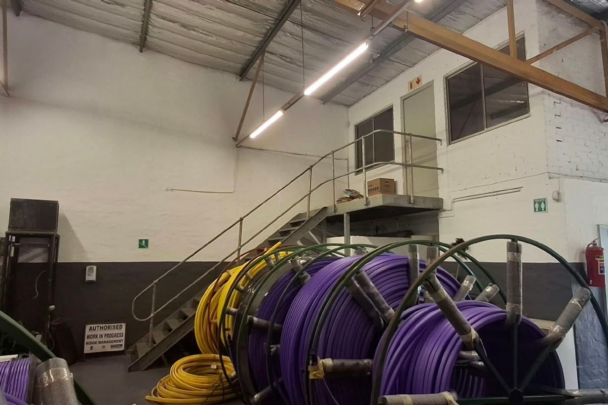 Industrial warehouse space featuring a mezzanine level with metal staircase access, exposed steel roof trusses, and functional overhead lighting.