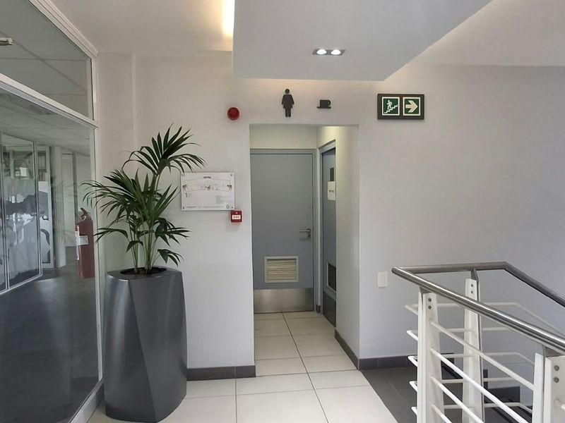 A clean, modern office hallway featuring glass partitions, tiled flooring, and clear emergency signage leading to restroom facilities.
