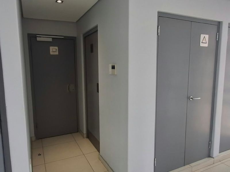 A clean, modern office hallway featuring tiled flooring, recessed lighting, and multiple access doors with clear safety and facility signage.