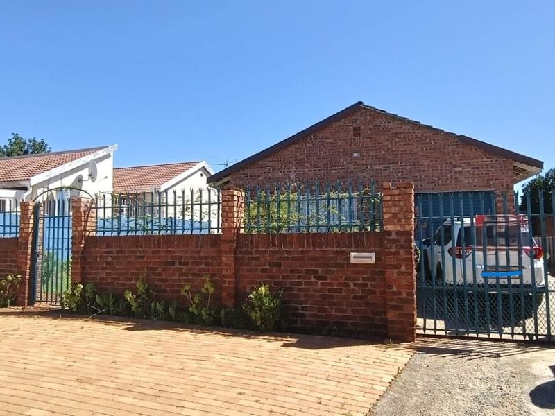 Front view of a face brick house featuring a secure gated entrance, paved driveway, and perimeter wall with metal fencing.