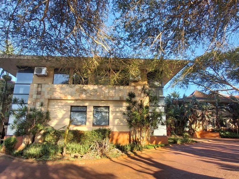 Front elevation of a commercial building featuring stone cladding, large windows, and a paved driveway surrounded by mature trees and landscaped gardens.