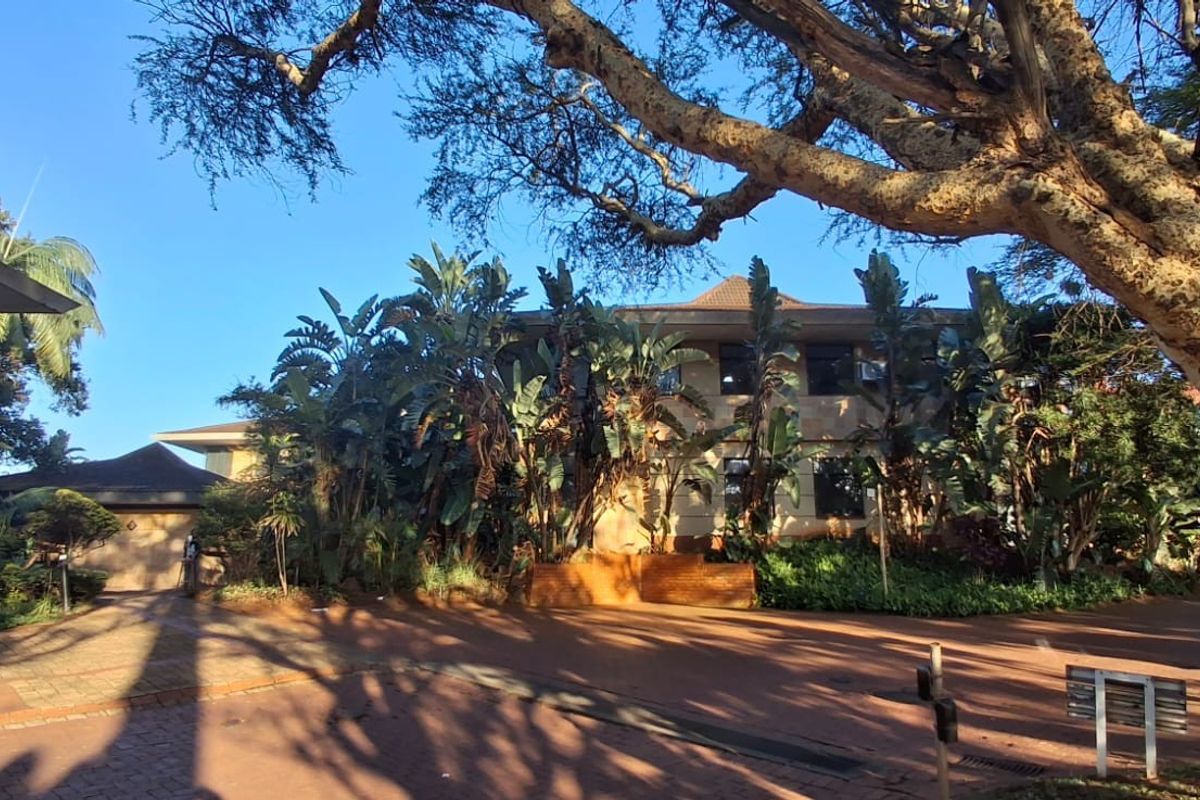 Front view of a multi-story commercial building featuring a paved driveway and established garden landscaping under mature trees.