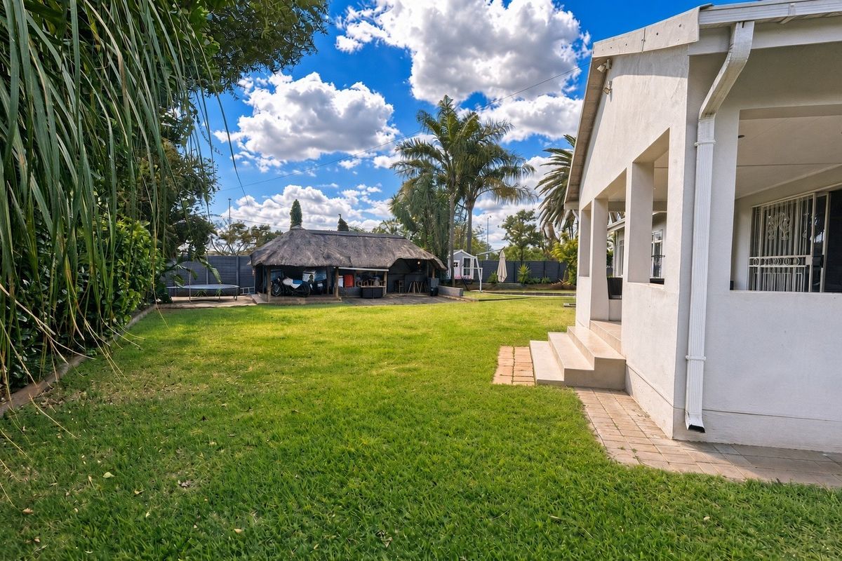 Spacious rear garden featuring a well-maintained lawn, a covered patio area, and a traditional thatched lapa structure.