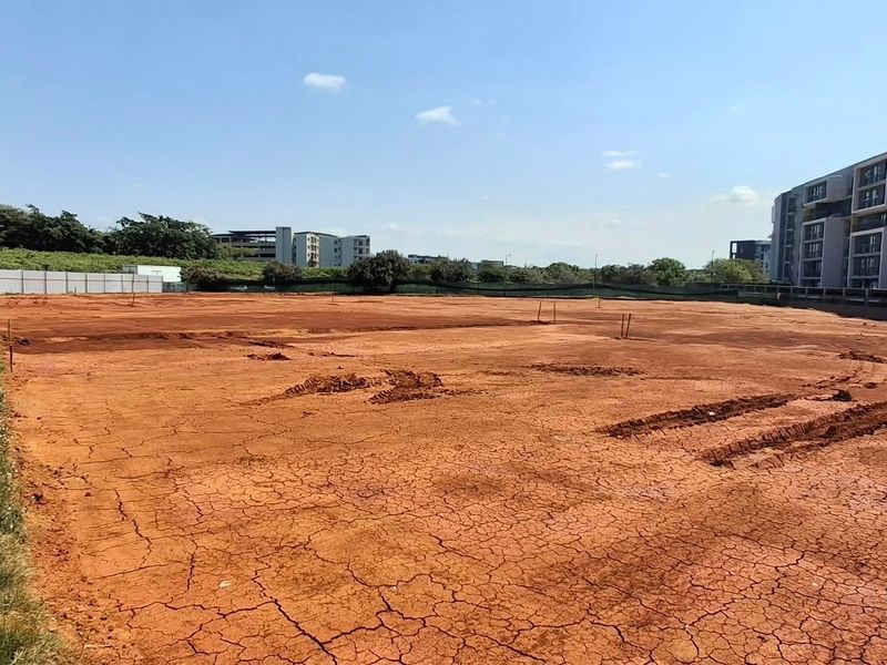 A vacant commercial land plot featuring cleared red earth and perimeter wooden fencing, situated adjacent to a modern multi-story residential complex.