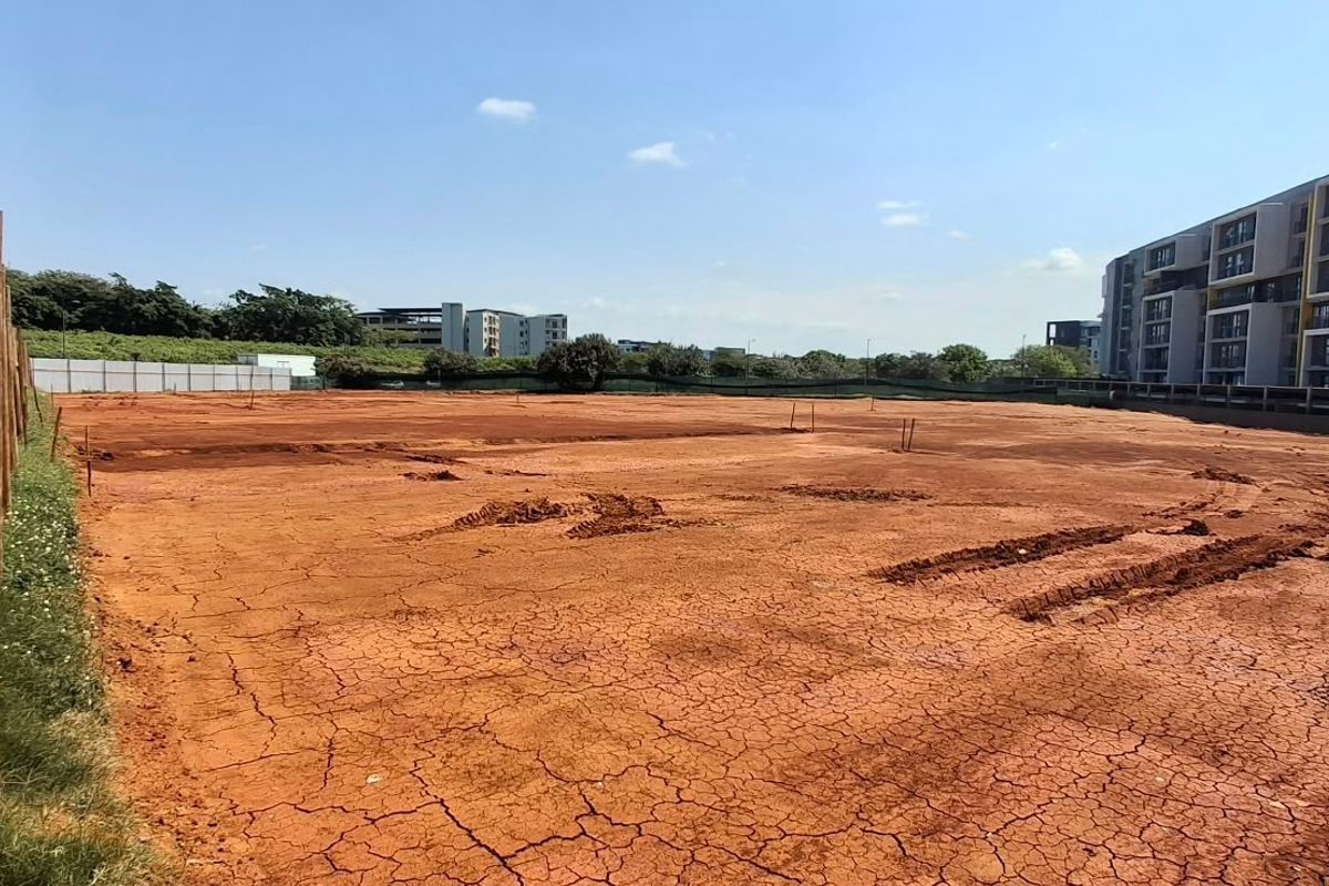 A vacant commercial land plot featuring cleared red earth and perimeter wooden fencing, situated adjacent to a modern multi-story residential complex.