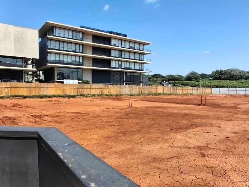A view of a cleared, vacant commercial plot of land with perimeter fencing, situated in an urban area near modern multi-story buildings.