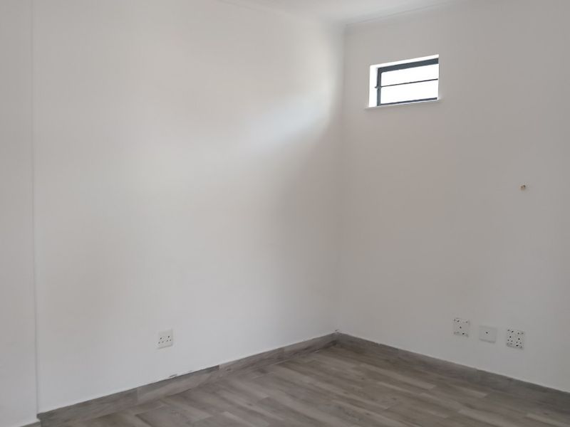 A clean, empty office space featuring modern laminate flooring, neutral white walls, and a small high-level window for natural light.