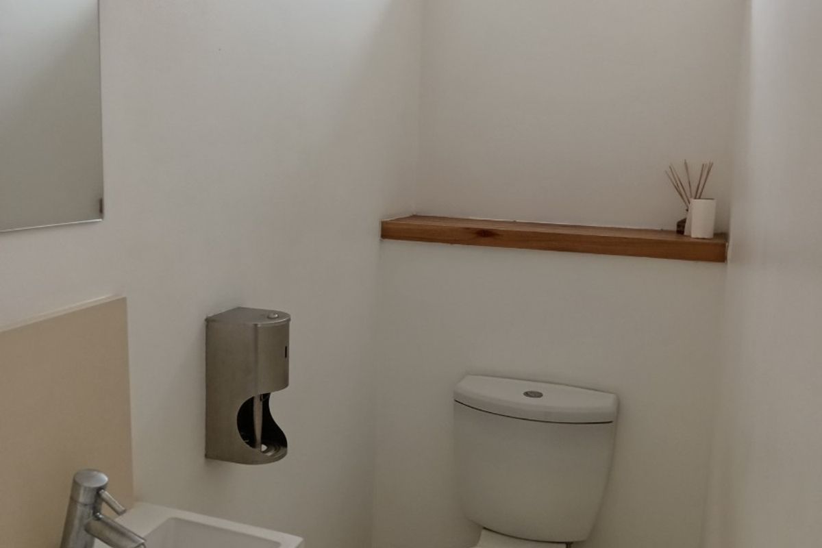 A clean and functional office restroom featuring a wall-mounted basin, toilet, tiled flooring, and a high-level window for natural light.