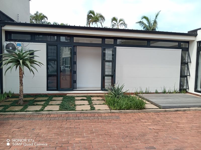 Modern office exterior featuring clean white walls, dark aluminium framed glass doors, timber decking, and a paved driveway area.