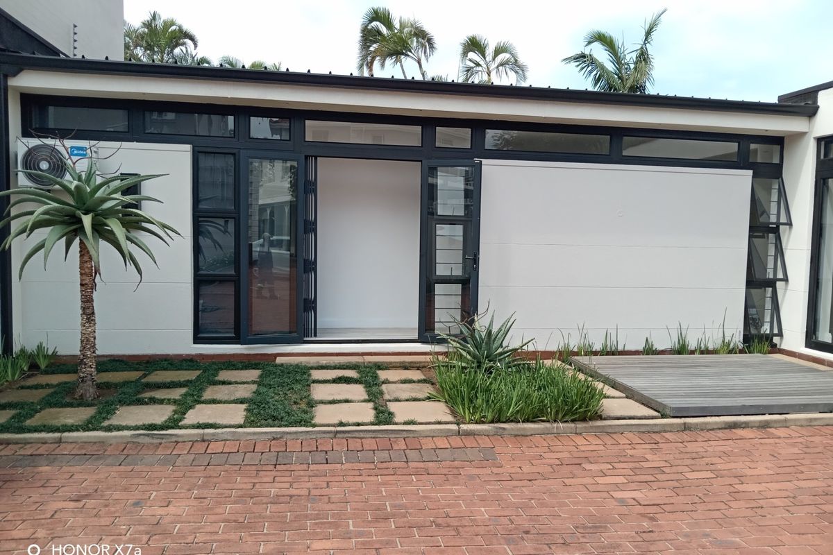 Modern office exterior featuring clean white walls, dark aluminium framed glass doors, timber decking, and a paved driveway area.