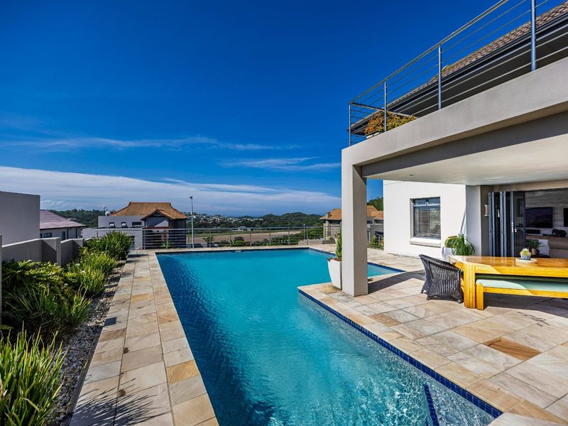 A modern outdoor pool area featuring a tiled deck, covered patio space, and elevated balcony with stainless steel railings, offering scenic views.