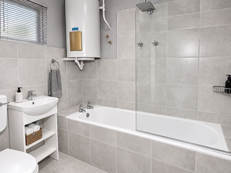 Modern bathroom featuring a bathtub with a glass shower screen, pedestal basin, toilet, and a wall-mounted electric water heater with neutral grey tiling.