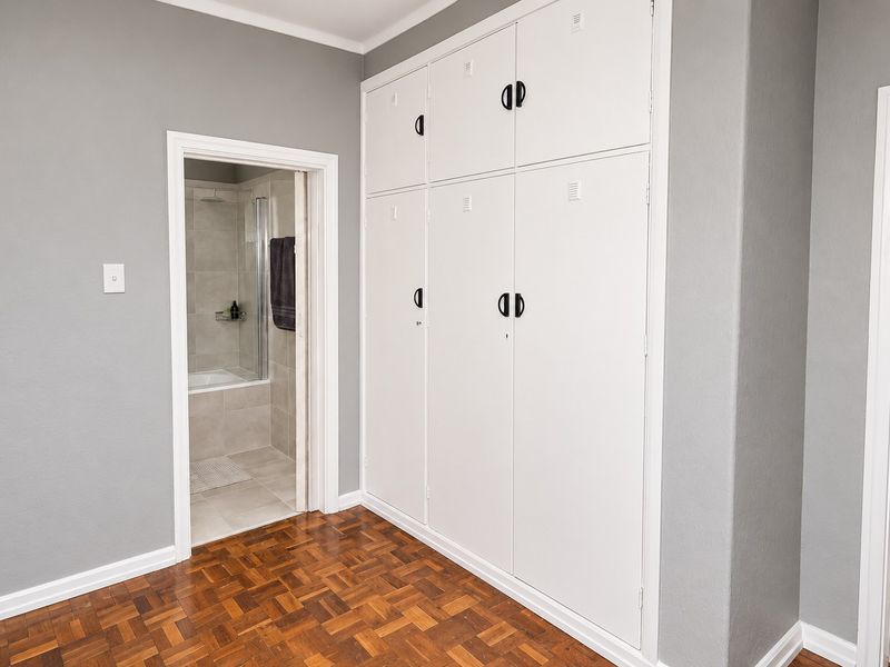 A clean bedroom featuring classic parquet flooring and extensive floor-to-ceiling built-in cupboards, with direct access to an adjoining bathroom.