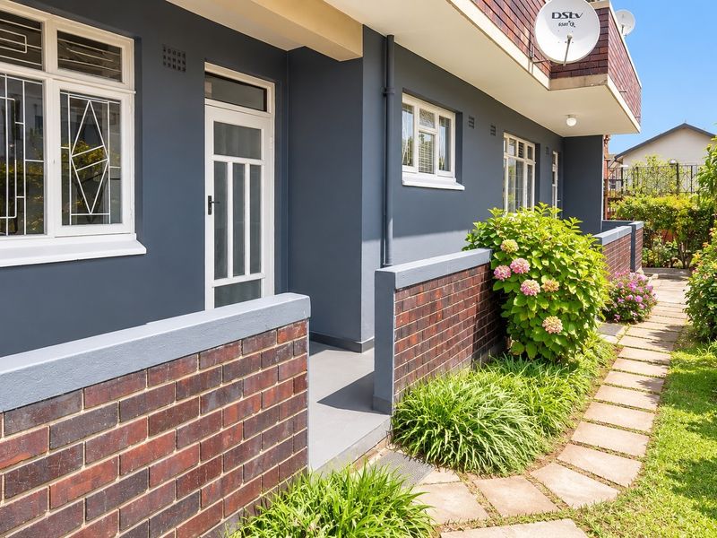 A well-maintained apartment exterior featuring a paved walkway, brick boundary walls, and secure window treatments, set against a landscaped garden area.