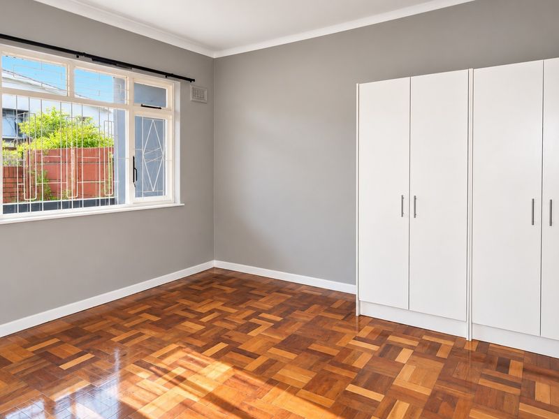 A bright, empty bedroom featuring polished parquet flooring, neutral grey walls, and white built-in cupboards with modern hardware.