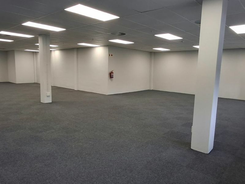 A spacious, open-plan commercial office area featuring carpeted flooring, suspended ceilings with recessed lighting, and large windows providing natural light.