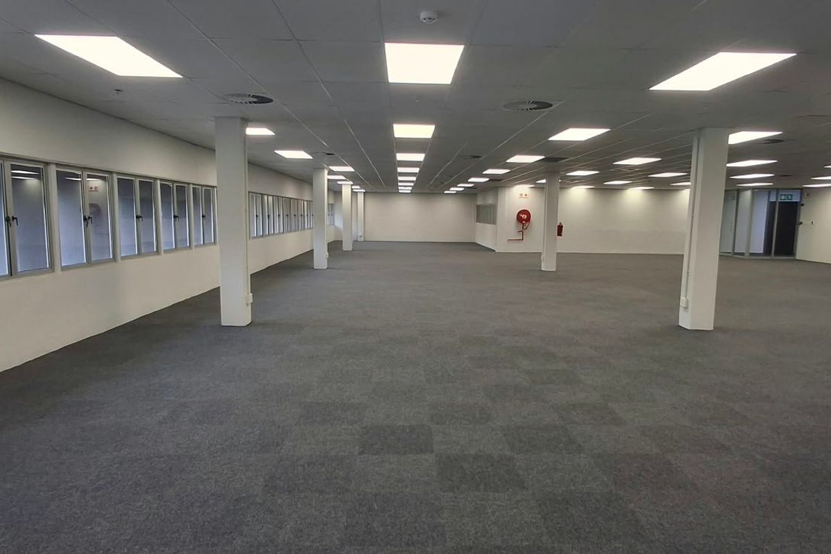 A spacious, open-plan commercial office area featuring a suspended ceiling, recessed lighting, carpeted flooring, and large windows providing natural light.