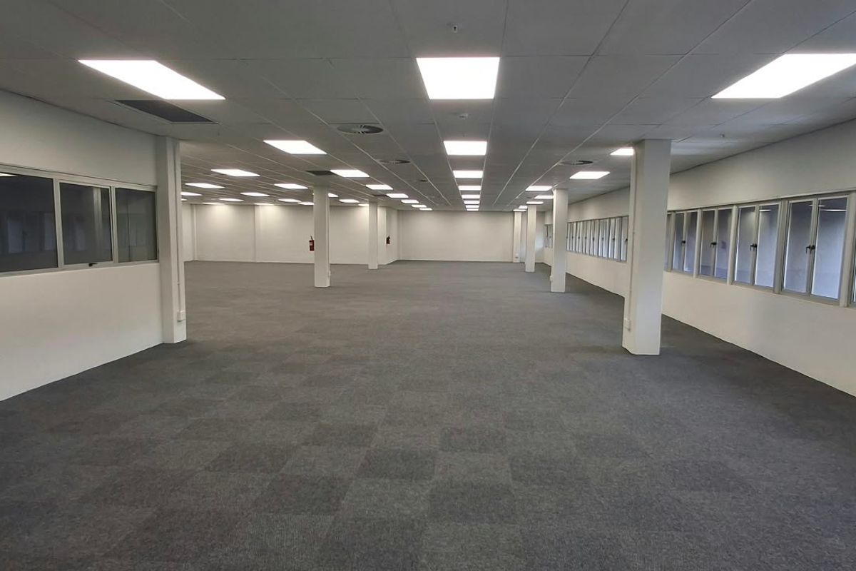 A spacious, open-plan commercial office area featuring neutral carpet tiles, suspended ceilings with recessed lighting, and large windows providing ample natural light.