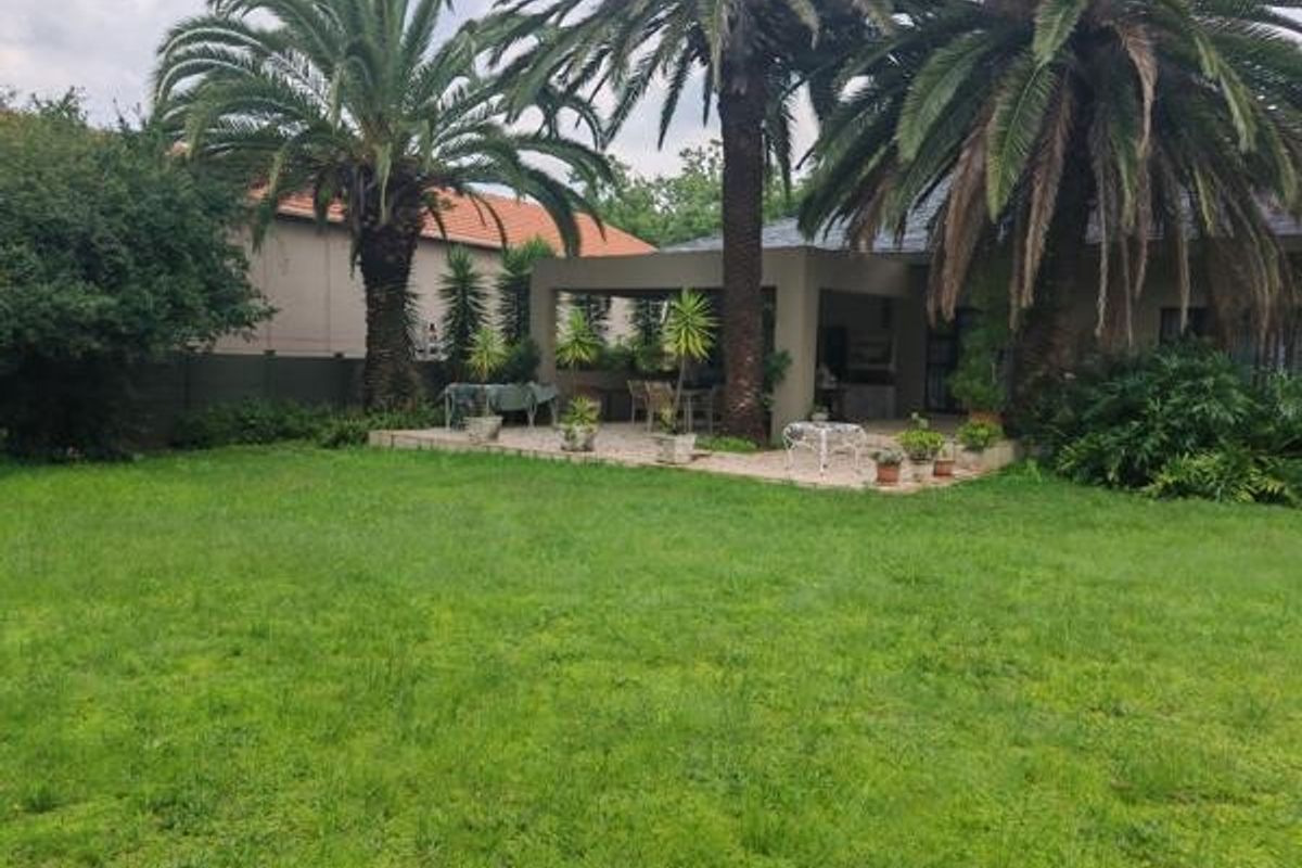 A spacious rear garden featuring a covered patio area, paved terrace, and mature palm trees, providing a private outdoor entertainment space.